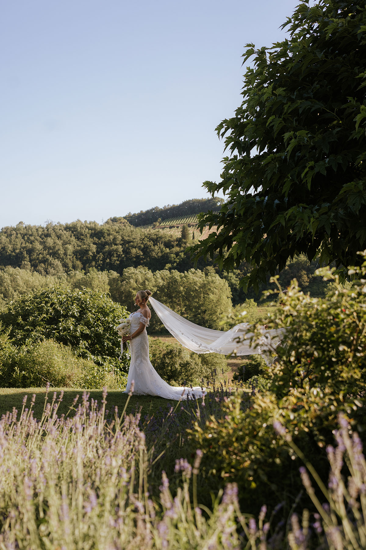 Bride in a fitted lace gown holding a bouquet, standing in a Tuscan garden with rolling hills behind her as her long veil flows in the breeze