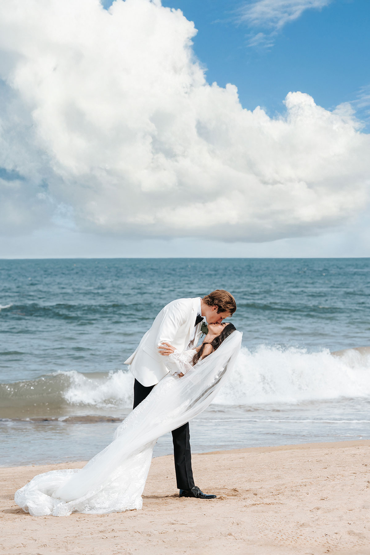 Bride and groom kissing on a sandy Scottish beach, veil flowing in the sea breeze