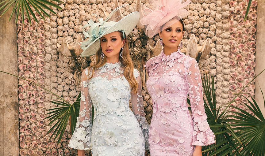 Two women wearing pastel lace occasionwear dresses with matching statement hats, posed against a textured backdrop.