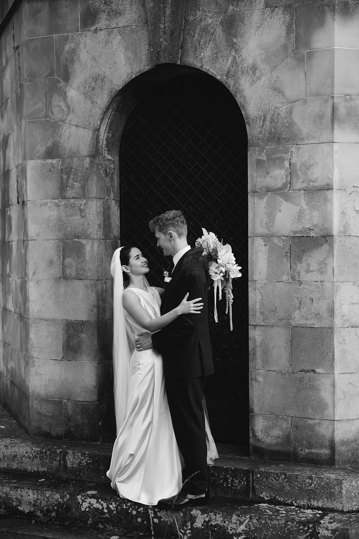 Black and white photo of bride and groom embracing outside a stone archway.