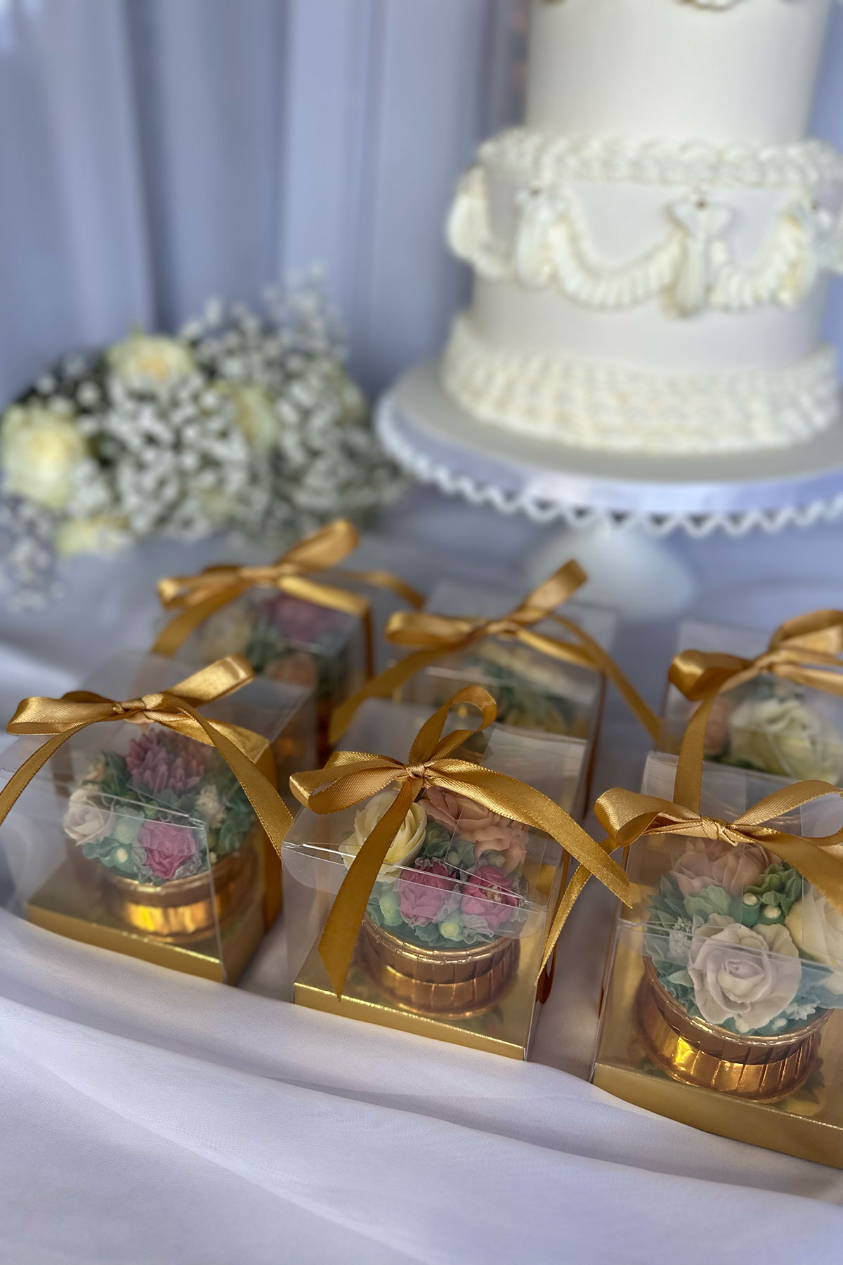 Boxed floral cupcakes with gold ribbon displayed beside a white wedding cake as edible wedding favours