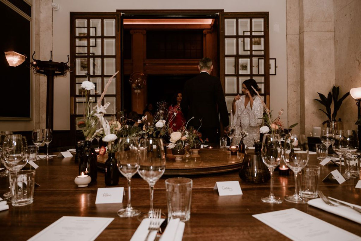Couple entering a wedding reception room with long wooden table, candles and floral table styling