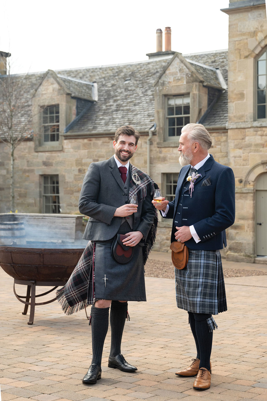 two men wearing kilt outfits from Eight Yards standing in the courtyard at Rosebery Steading in front of a fire pit