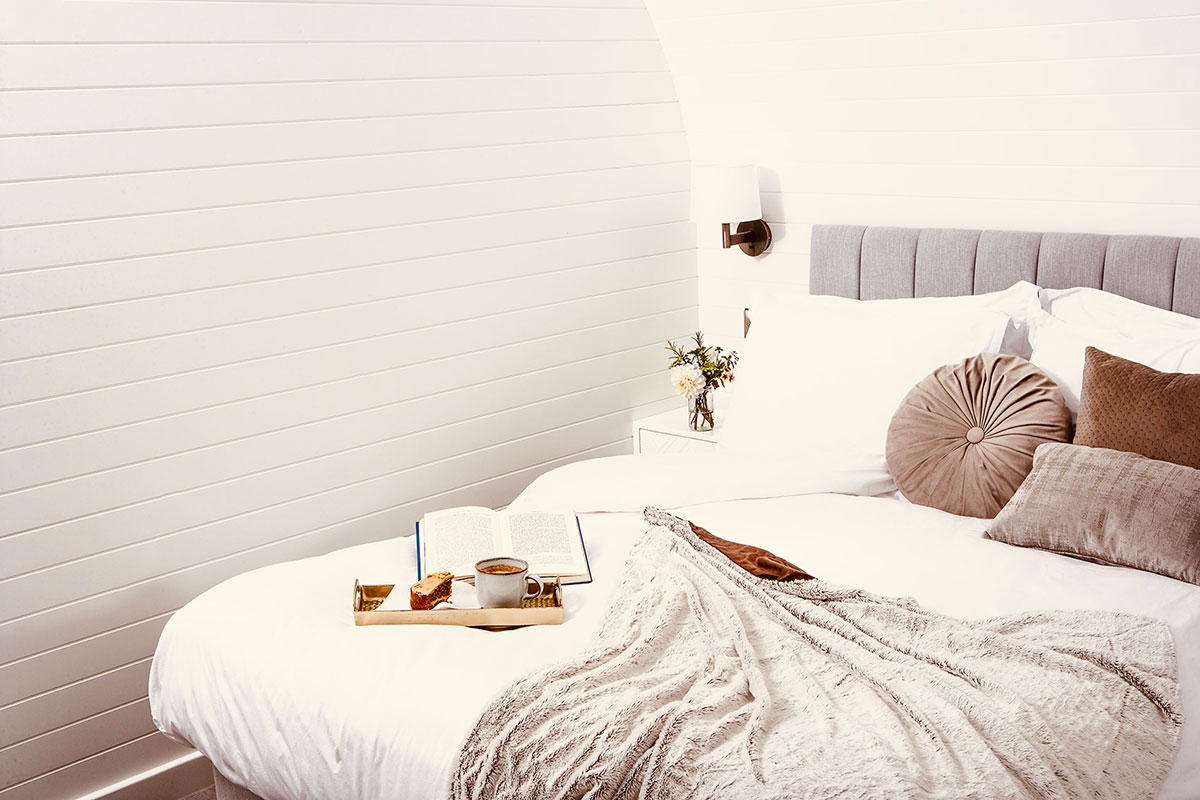 a white sheeted bed with brown pillows, a grey blanket, an open book and a tray with coffee and cake sat on top, situated in the corner of a white panelled room