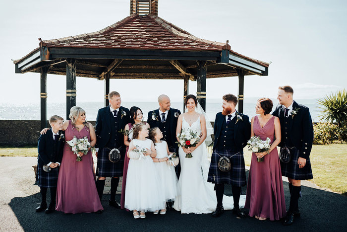 a group portrait of bride, groom, bridesmaids and groomsmen taken under a wooden pagoda outside on the seafront at Seamill Hydro