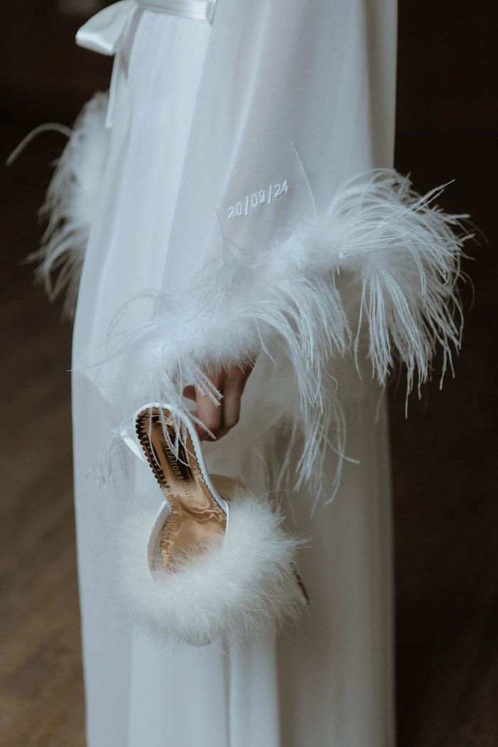 A close up of a bride wearing a white robe with fluffy feather trim on the sleeves and the date 20/09/24 embroidered on the sleeve. She is holding a pair of white shoes with feathers across the front strap