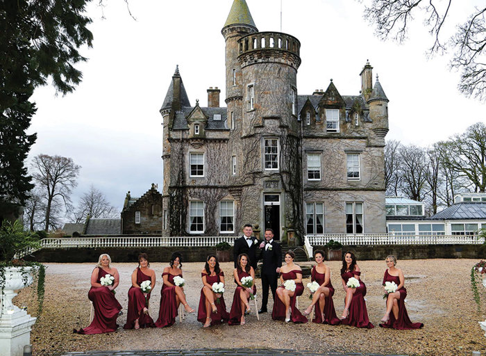 nine bridesmaids wearing burgundy dresses pose seated as two grooms wearing smart black suits and bow ties stand behind them. The exterior of Carlowrie Castle is in the background