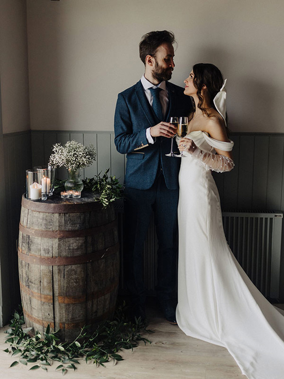 bride and groom cheers champagne glasses stood next to wooden barrel with candles and greenery on top