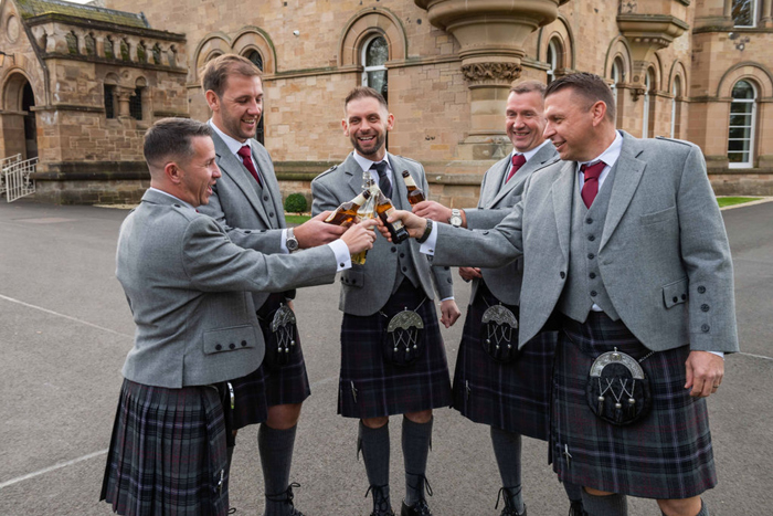Five Men Wearing Kilts And Grey Jackets Holding Beer Bottles Outside A Castle