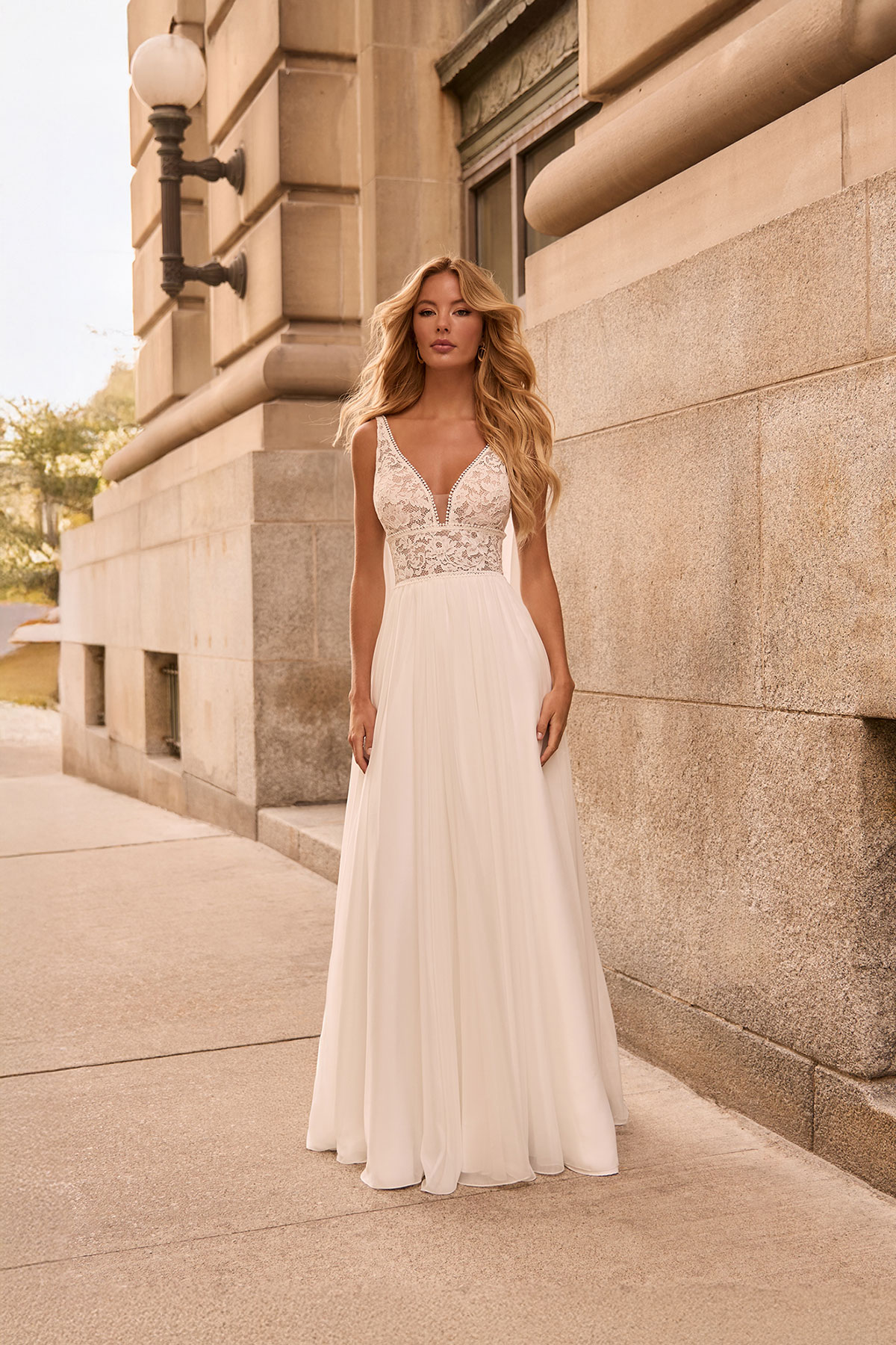 Bride in a simple ivory wedding dress with a lace V-neck bodice, sheer detailing and a softly flowing skirt, standing outdoors against a stone building