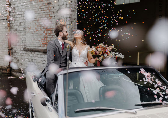 bride and groom sit in cream coloured convertible car wearing heart shaped sunglasses and surrounded by colourful falling confetti