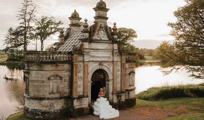 A bride and groom standing in front of an ornate small Victorian building at Kinmount House