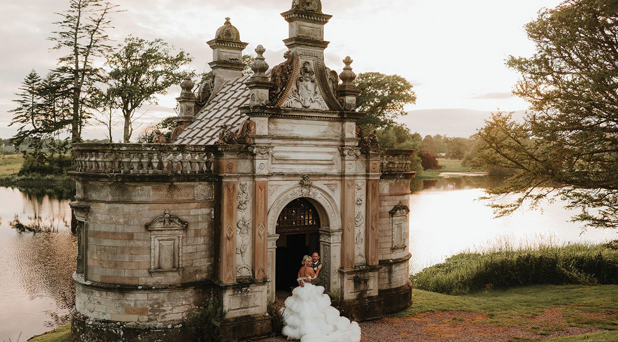 A bride and groom standing in front of an ornate small Victorian building at Kinmount House