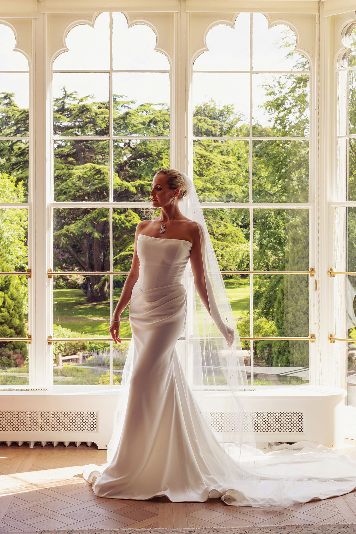 Bride wearing a fitted strapless wedding dress and long veil in front of large arched windows.
