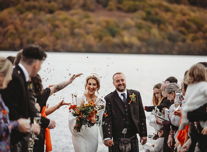 a bride and groom walk through two rows of guests as they throw confetti at them. There is a loch and hills in the background