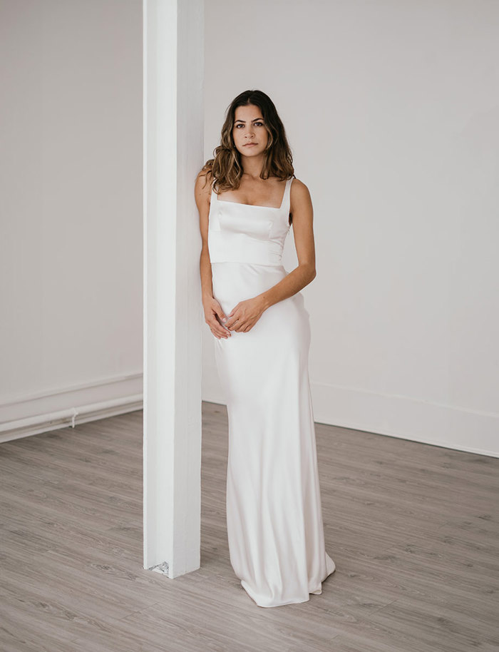 a brunette stands wearing a full-length silky white gown with her arms meeting in front of her waist