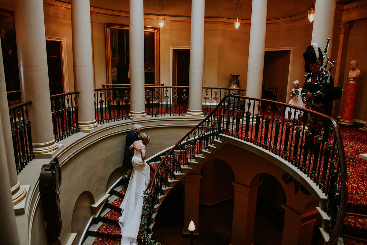 A bride and piper on the grand curved staircase inside Culzean Castle, with red carpet and classical columns.
