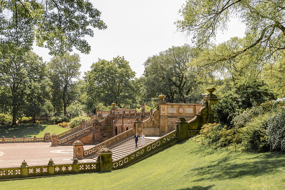 Wide shot of a couple in wedding attire walking down ornate stone steps surrounded by greenery in Central Park