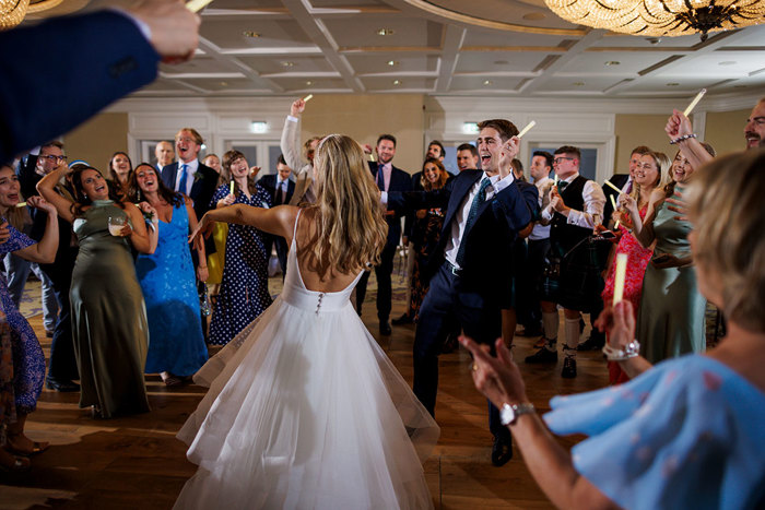 A joyful bride and groom dance enthusiastically at their Old Course Hotel wedding, surrounded by cheering friends and family in a lively reception hall
