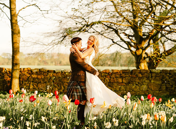 a groom lifts a joyous bride up. They are in a garden that's carpeted with colourful tulips and small daffodils with a stone wall and trees in the background
