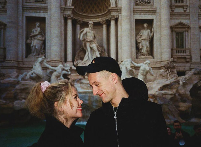 A woman and a man wearing black hoodies looking at each other smiling while standing in front of an ornate fountain
