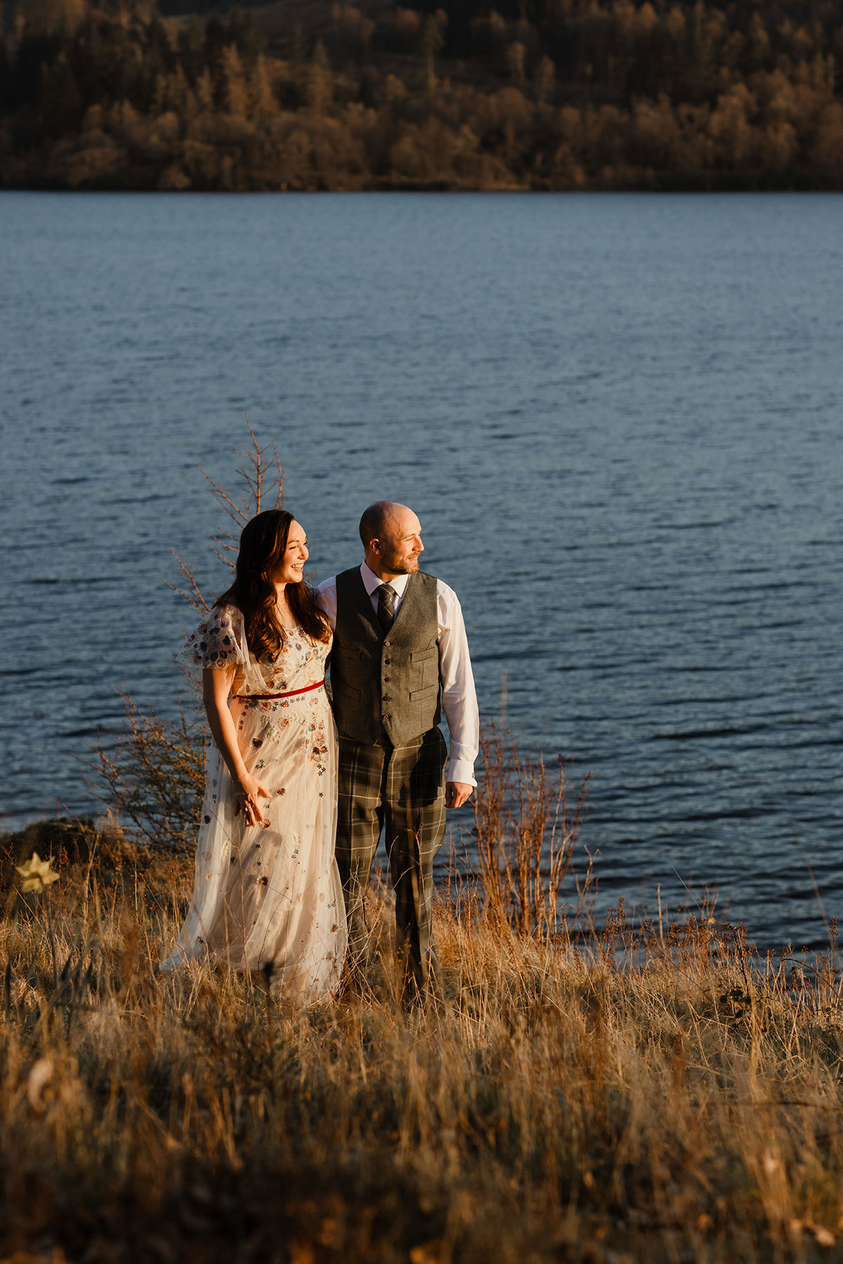Bride and groom standing by the loch at sunset