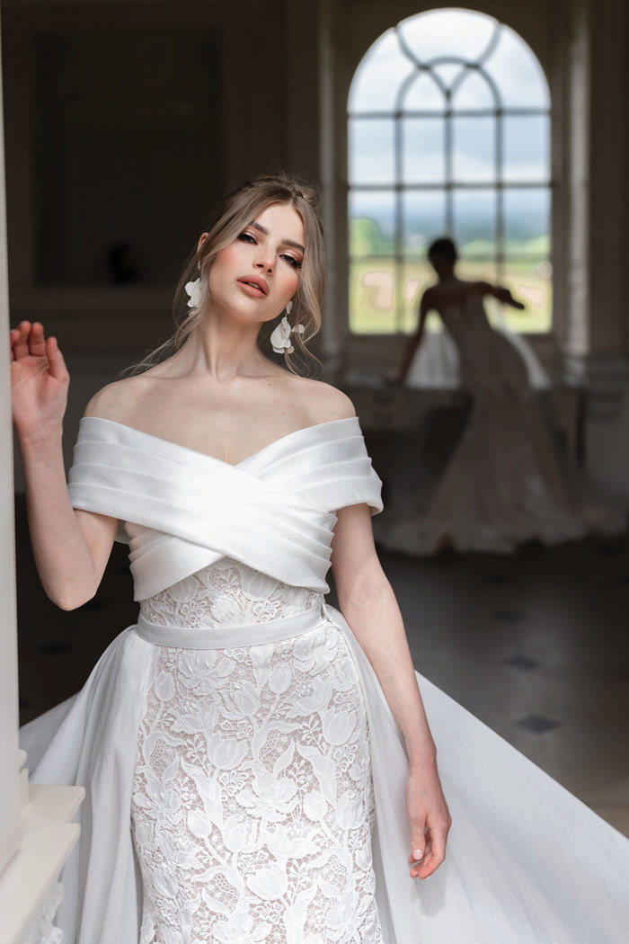 bride in off the shoulder wedding dress stands in front while another bride can be seen in the darkened back of the shot looking out the window