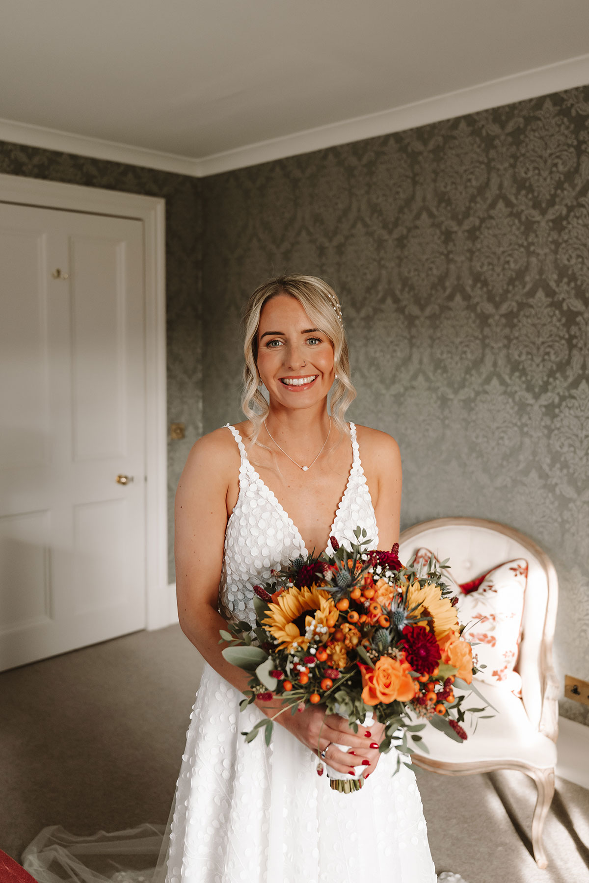Bride in white gown holding autumnal bouquet of sunflowers, roses, and berries inside Ballogie House.