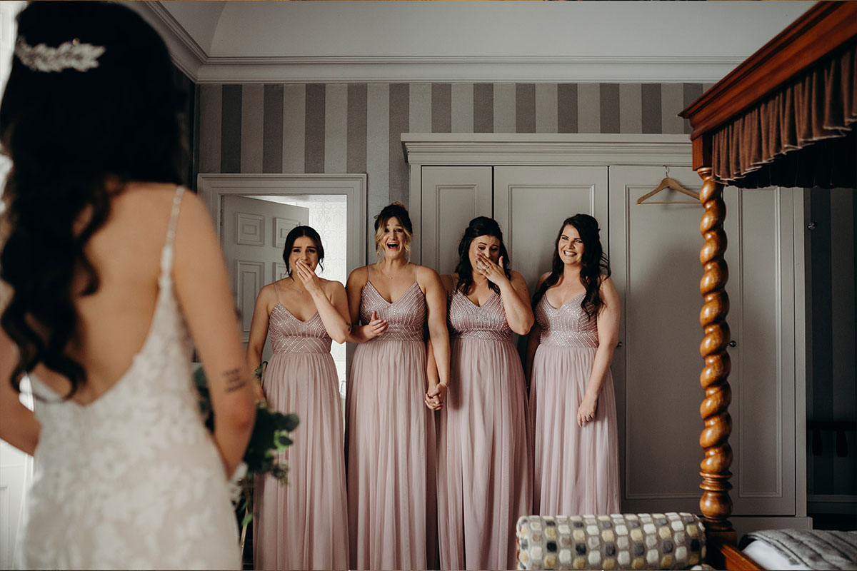 Four bridesmaids wearing pale dusky pink react to seeing the bride