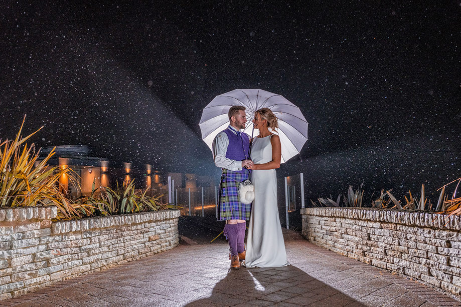 A bride and groom at nighttime under an umbrella outside The Vu wedding venue Scotland