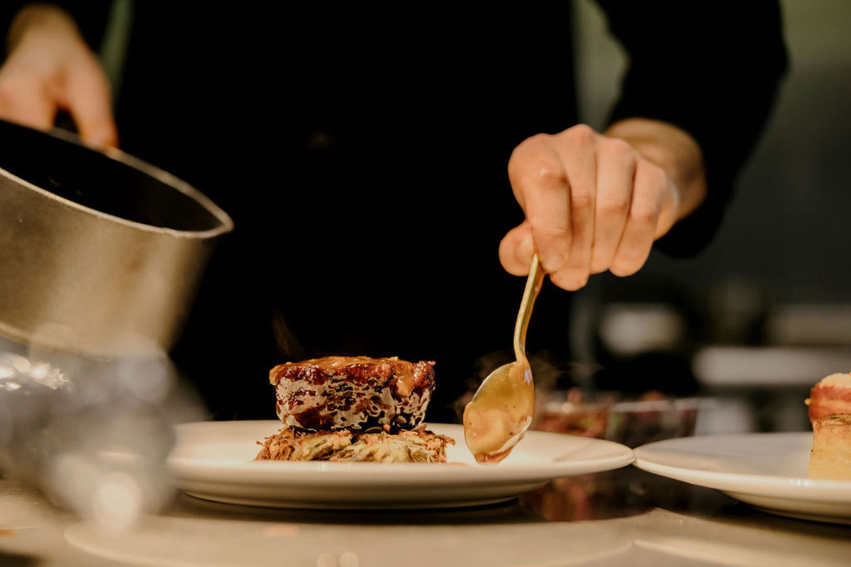 A chef plating a seared meat dish, spooning sauce onto a white plate in a professional kitchen