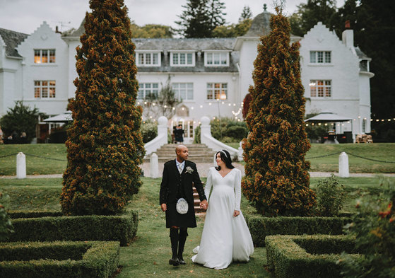 A person in a kilt and person in a wedding dress holding hands and walking in a garden at Achnagairn Castle.