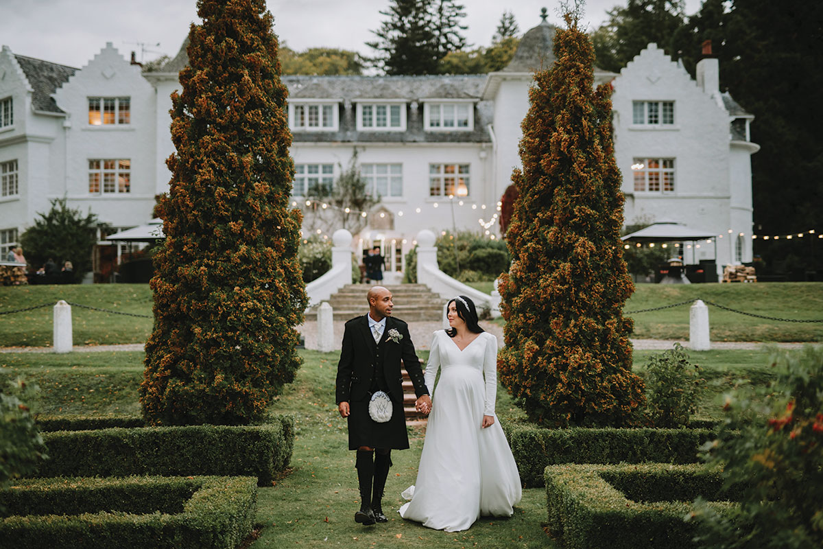 A person in a kilt and person in a wedding dress holding hands and walking in a garden at Achnagairn Castle.