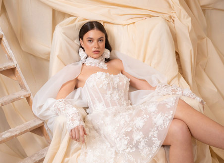 bride with dark brown hair wears strapless floral applique mini wedding dress and long matching sleeves as she sits in a chair covered by a dark cream sheet doubling as the backdrop
