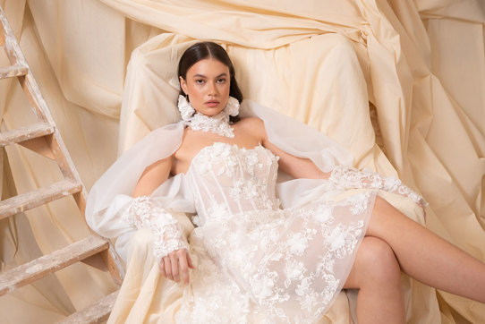 bride with dark brown hair wears strapless floral applique mini wedding dress and long matching sleeves as she sits in a chair covered by a dark cream sheet doubling as the backdrop