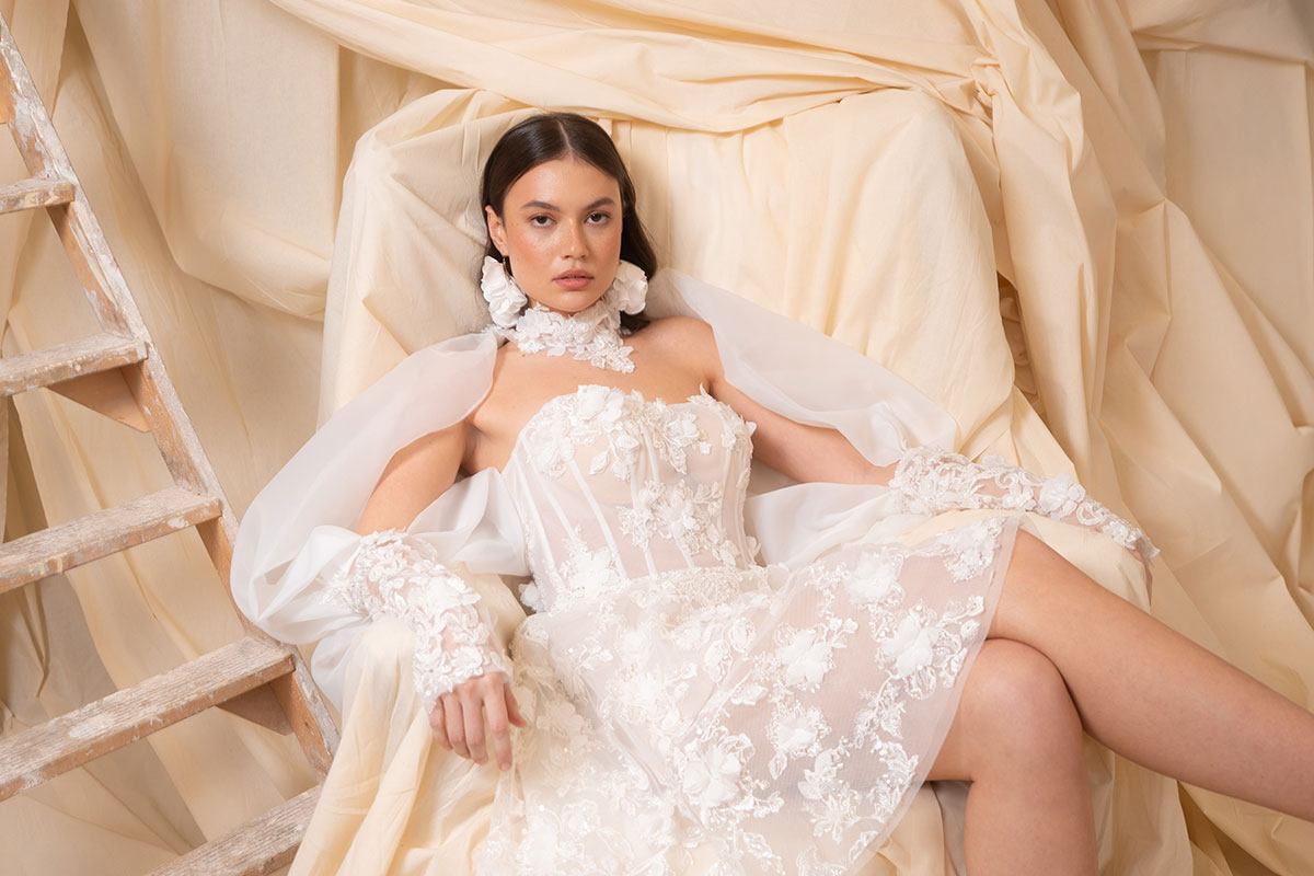 bride with dark brown hair wears strapless floral applique mini wedding dress and long matching sleeves as she sits in a chair covered by a dark cream sheet doubling as the backdrop