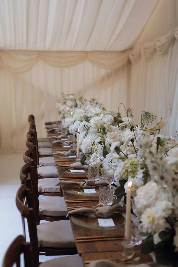 long wooden table set with clear charger plates, linen napkins, lit candlesticks and a long middle section filled with white flowers and crystal glassware sits nicely within a draped marquee