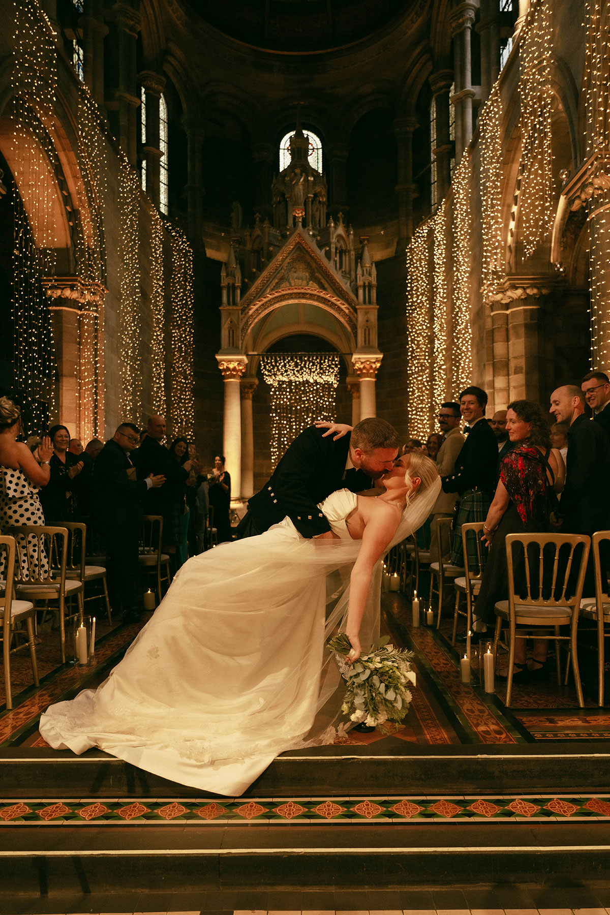 bride and groom dip kiss in aisle with fairy lights in grand historic wedding venue