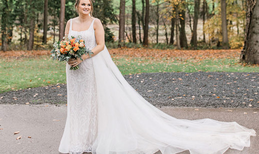 bride holding bouquet stands outside in sequinned wedding dress with long train and veil laid out to the side 