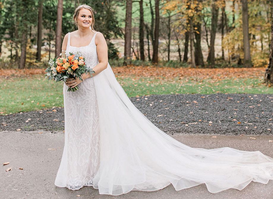 bride holding bouquet stands outside in sequinned wedding dress with long train and veil laid out to the side 