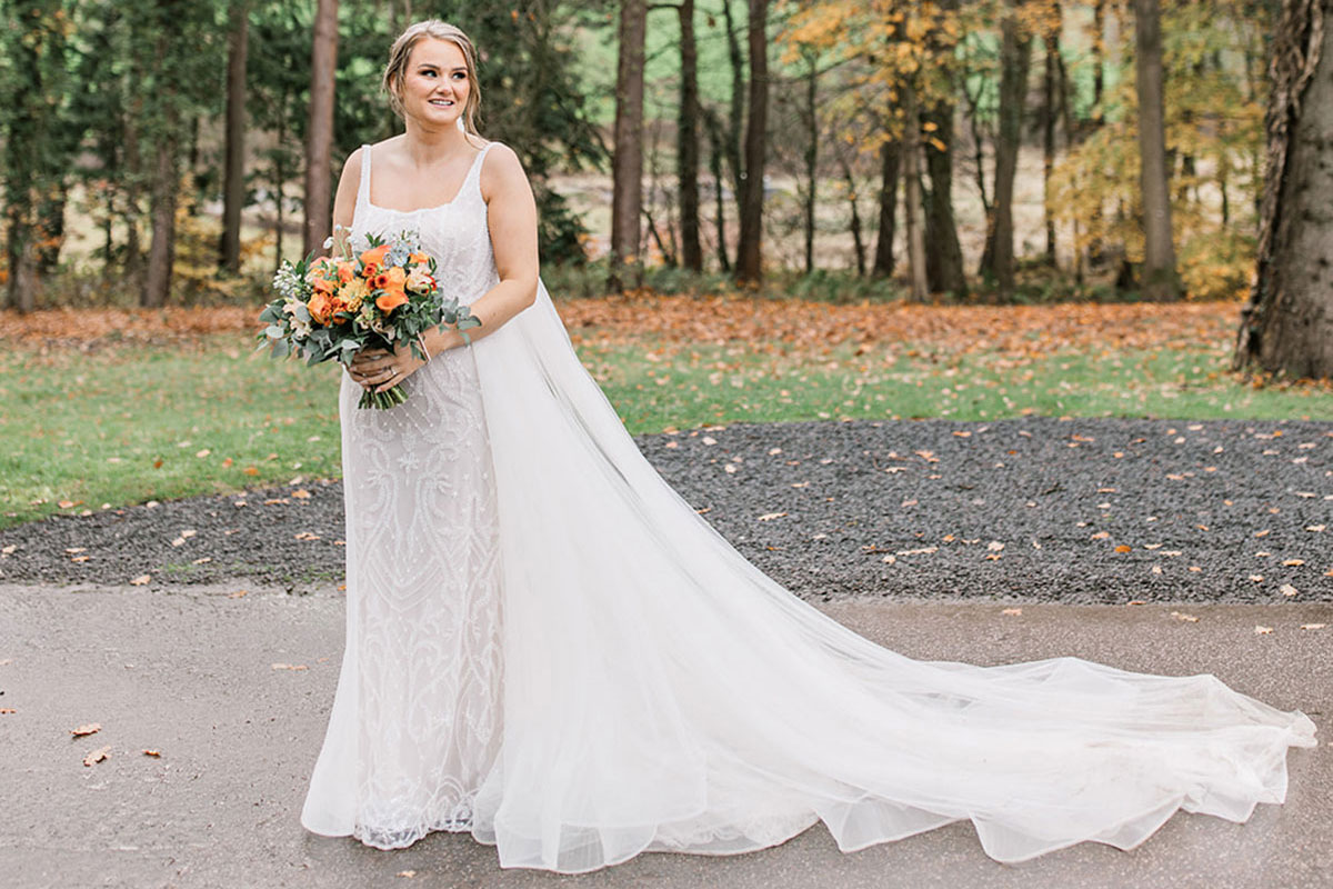 bride holding bouquet stands outside in sequinned wedding dress with long train and veil laid out to the side