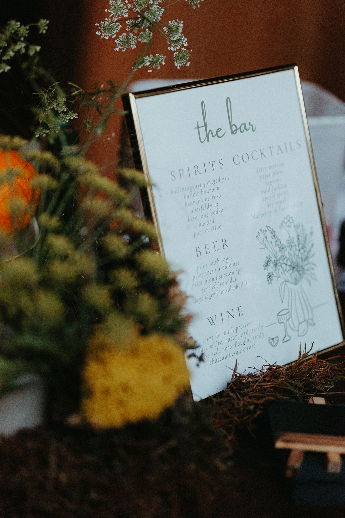 Illustrated wedding bar menu displayed among seasonal flowers and foliage at a Cambo Country House reception.