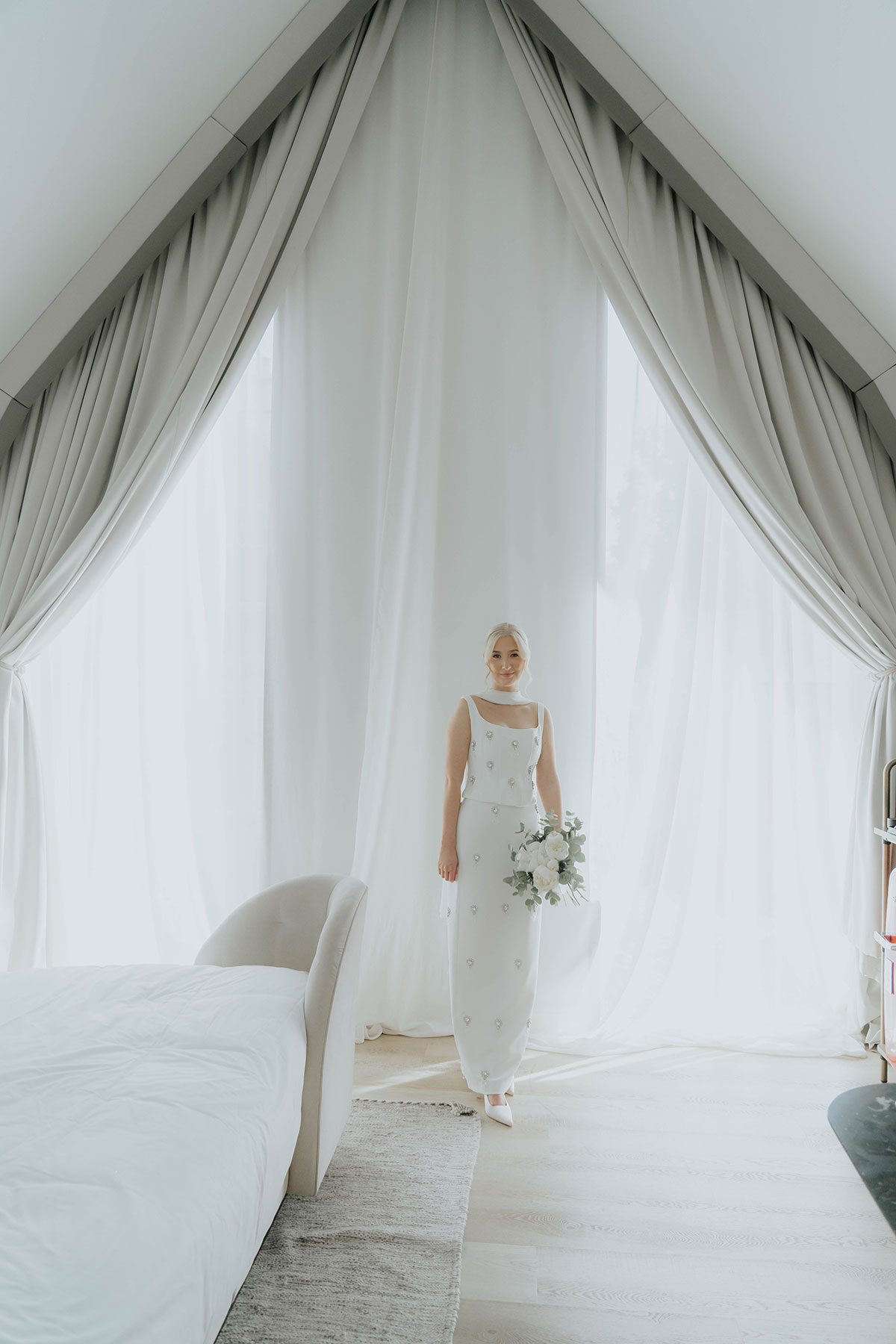 Bride in an embellished contemporary wedding dress holding a white bouquet in a bright modern hotel room