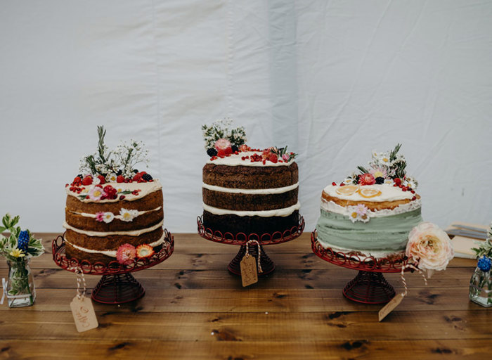 three cakes decorated with pretty flowers sitting on red wire cake stands on a wooden surface in a marquee