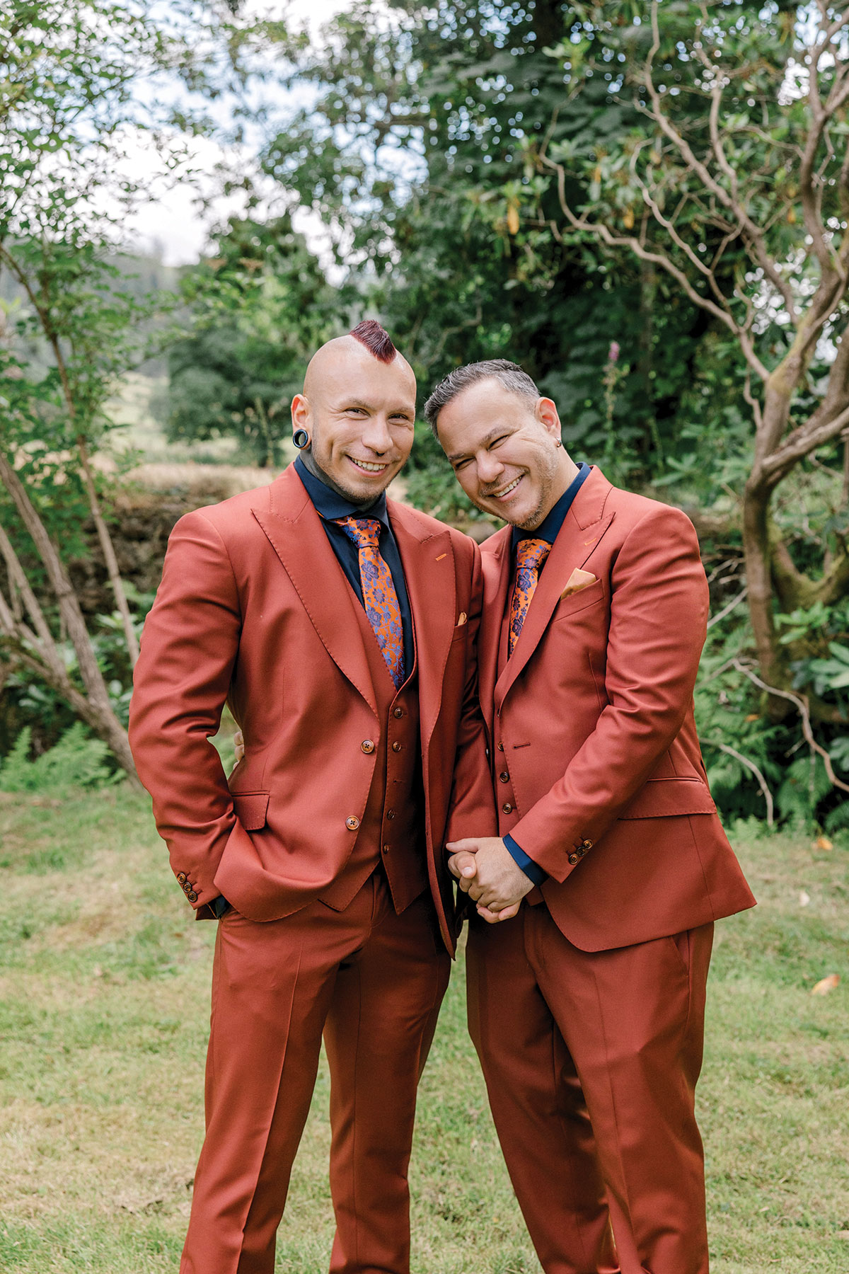 Two grooms wearing matching rust-coloured wedding suits posing outdoors at Venachar Lochside in Scotland