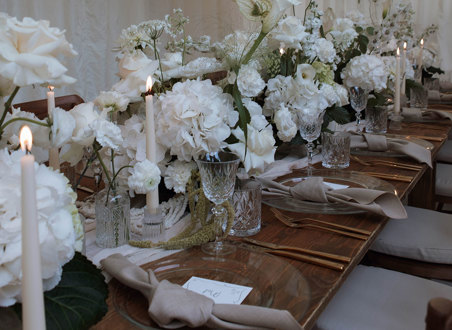 long wooden table set with clear charger plates, linen napkins, and a long middle section filled with white flowers and crystal glassware