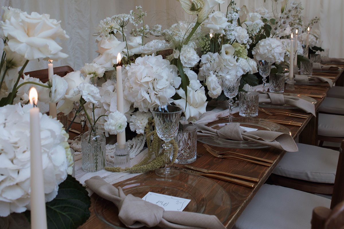 long wooden table set with clear charger plates, linen napkins, and a long middle section filled with white flowers and crystal glassware