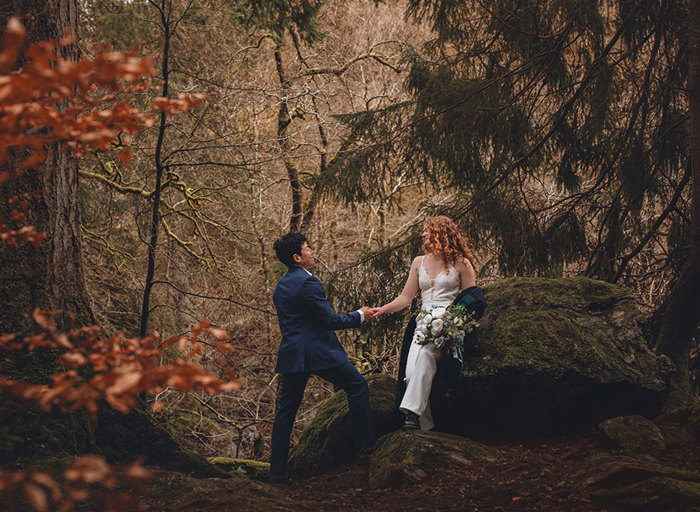 A bride in a blue suit and a bride in a white jumpsuit face each other holding hands in a forest