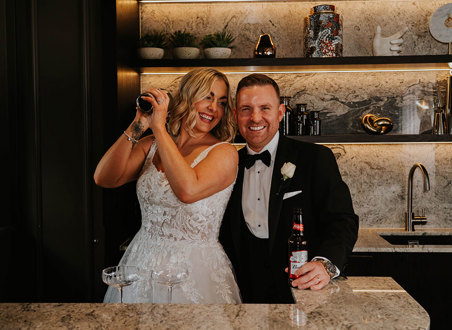 A bride and groom posing for a picture while making a cocktail.