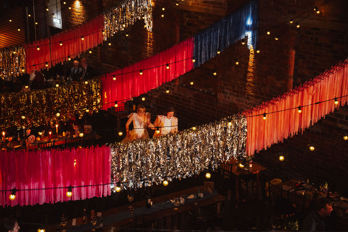 Wide shot of the Engine Works reception space with pink, blue and metallic streamers hung from the ceiling and warm festoon lighting, with the brides entering the room.